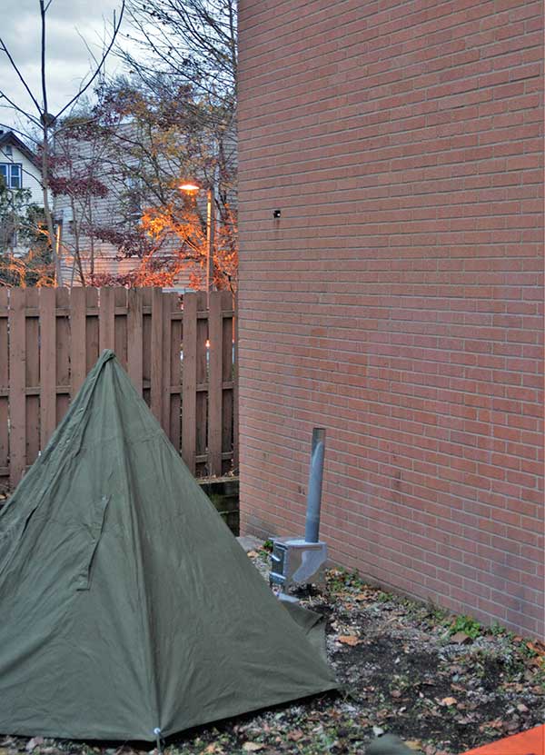 At dusk, author set up Polish Tarp Shelter with opening facing the brick wall and a woodburning stove. At night, shelter will be nearly impossible to see.